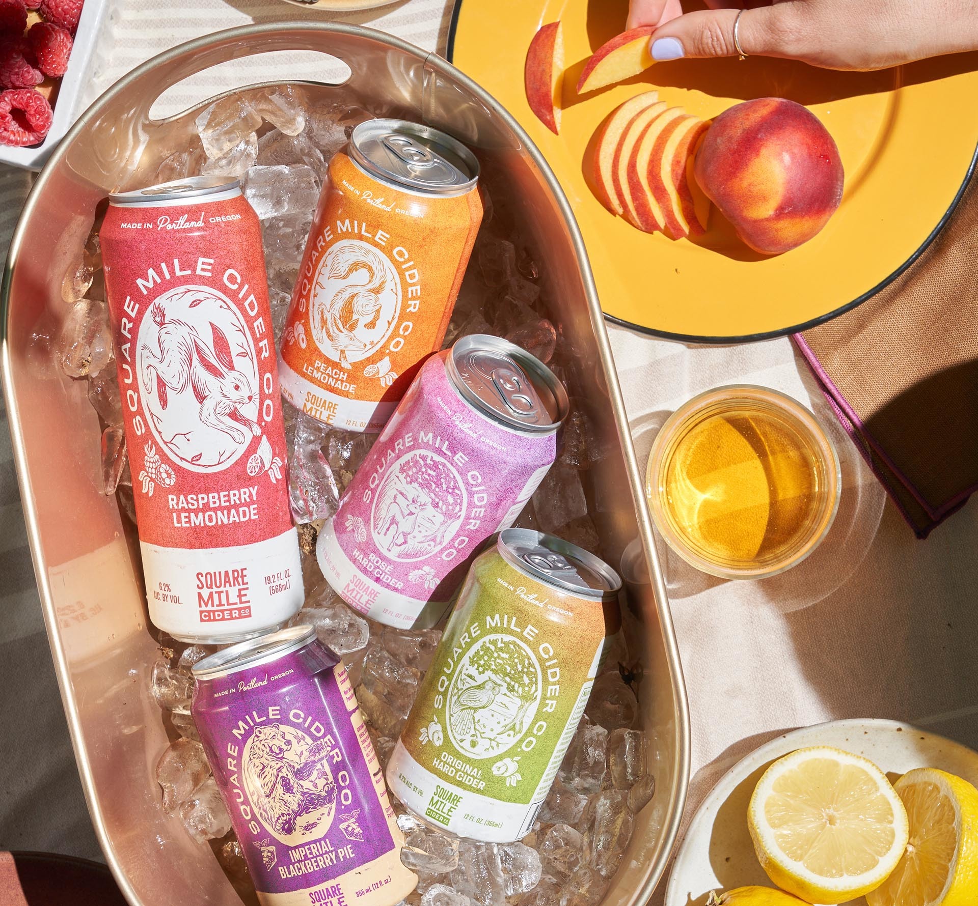 An overhead view of a big bucket filled with ice and cans of Square Mile Cider with fruit on plates on a table